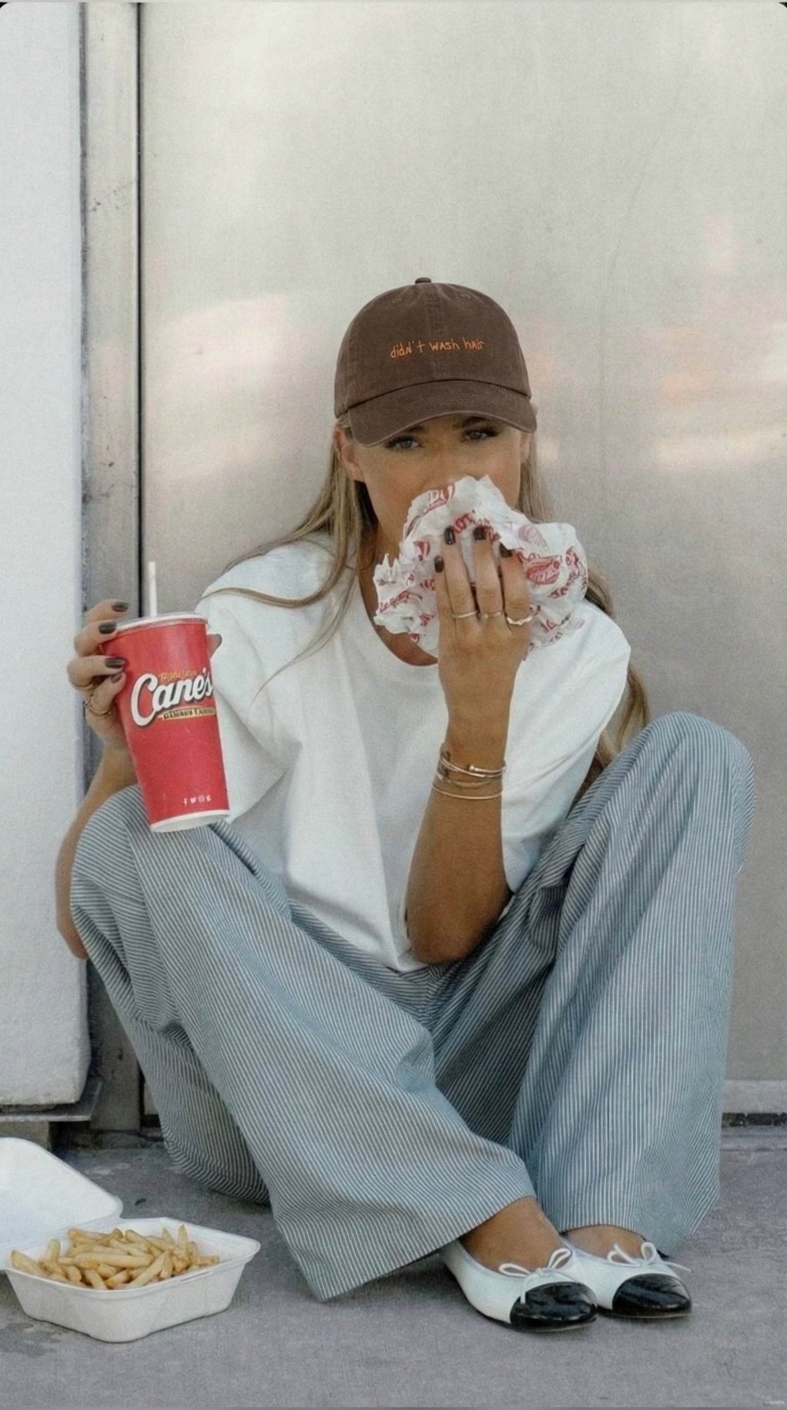 Female sitting on the ground holding a soft drink cup and eating fries and a burger, wearing a brown cap that’s says didn’t wash hair with wide  leg pants. 